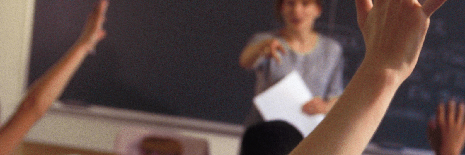 Being a Special Education Teacher During DeVos Era of Education Students raising their hands in classroom, with teacher at the front of the class in front of the chalkboard