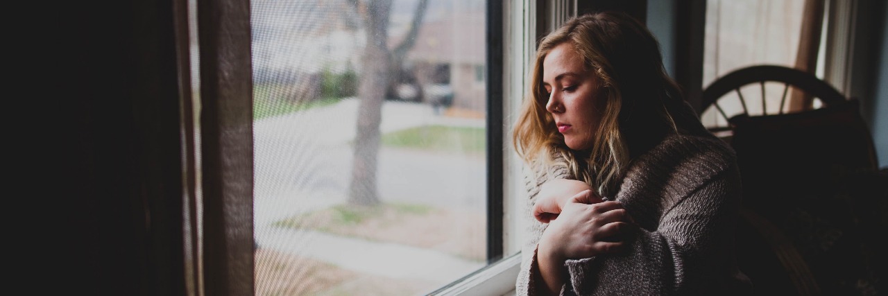 woman sitting beside window in darkened room looking out with sadness