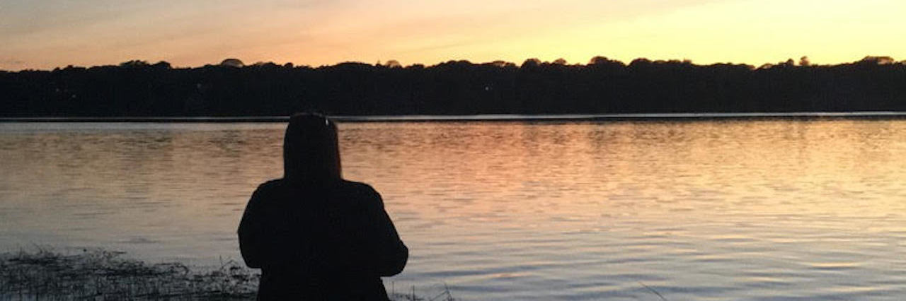Becoming a New Person With Lyme Disease woman standing at the edge of a lake looking at the sunset