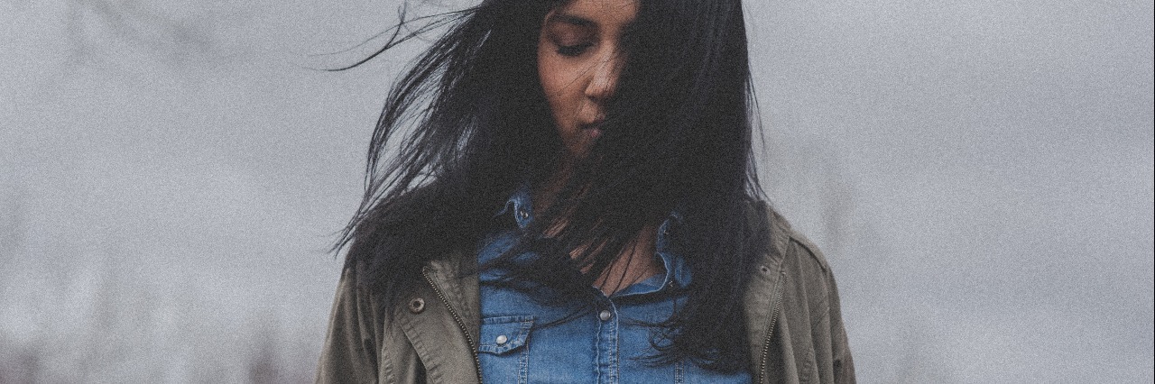 image of dark haired woman standing in front of cloudy sky with hair partly covering face