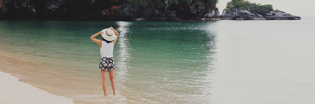 How Traveling Changes When You Have PTSD woman standing in ocean on idyllic beach with rock formation in distance