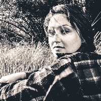 black and white image of woman in checkered shirt sitting in field looking at camera straight-faced