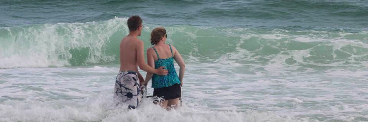The author standing in the ocean with her walker, with her son standing next to her