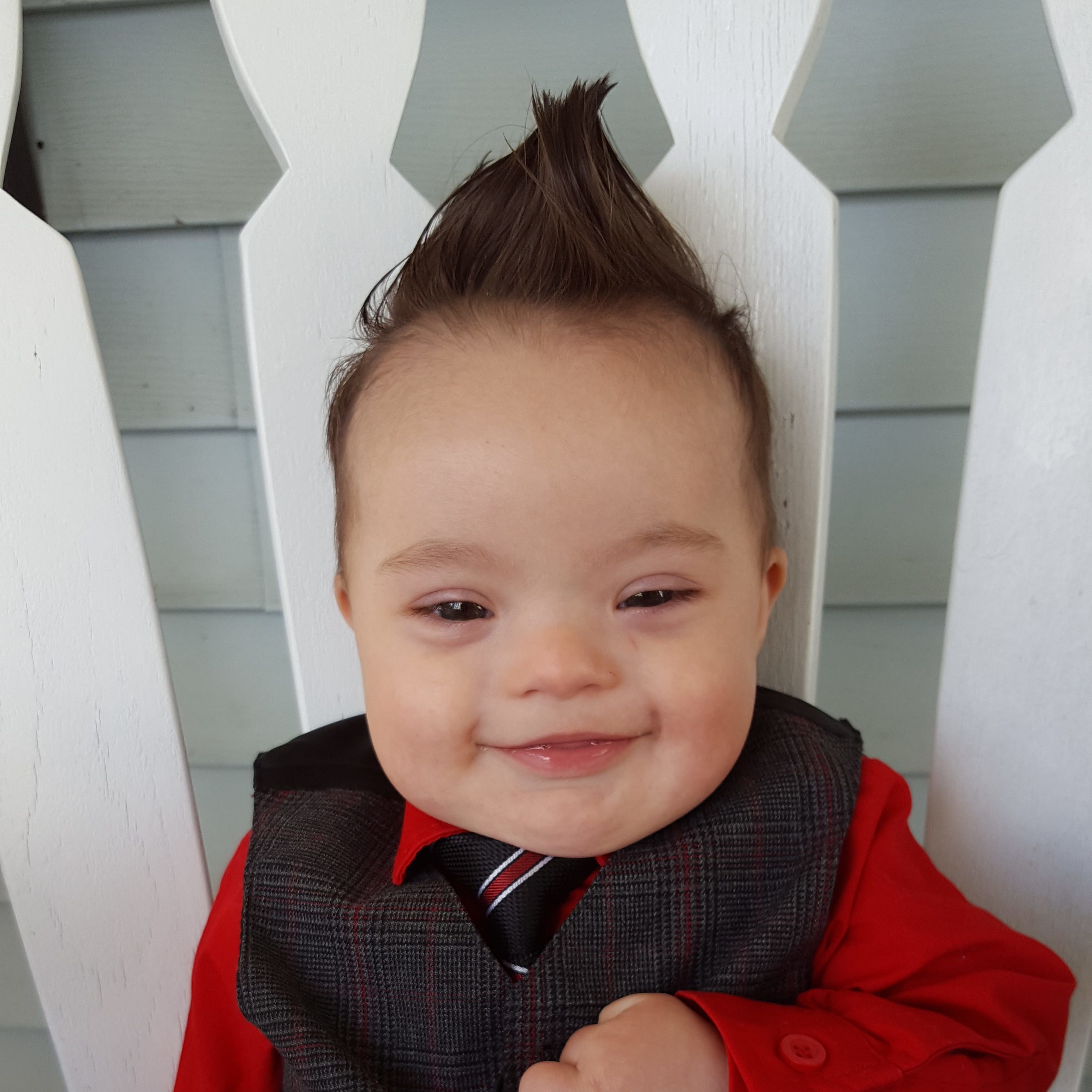 Little baby boy with Down syndrome smiling, his thicj dark hair is styled sticking up like a Mohawk and he is wearing a suit with a red shirt