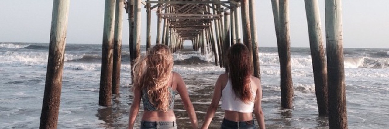 How to Help and Support a Loved One With a Chronic Illness two sisters holding hands under a pier at the beach