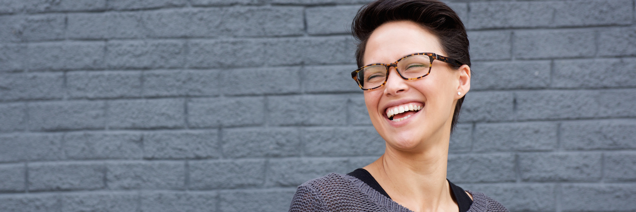 woman with glasses laughing in front of a gray wall