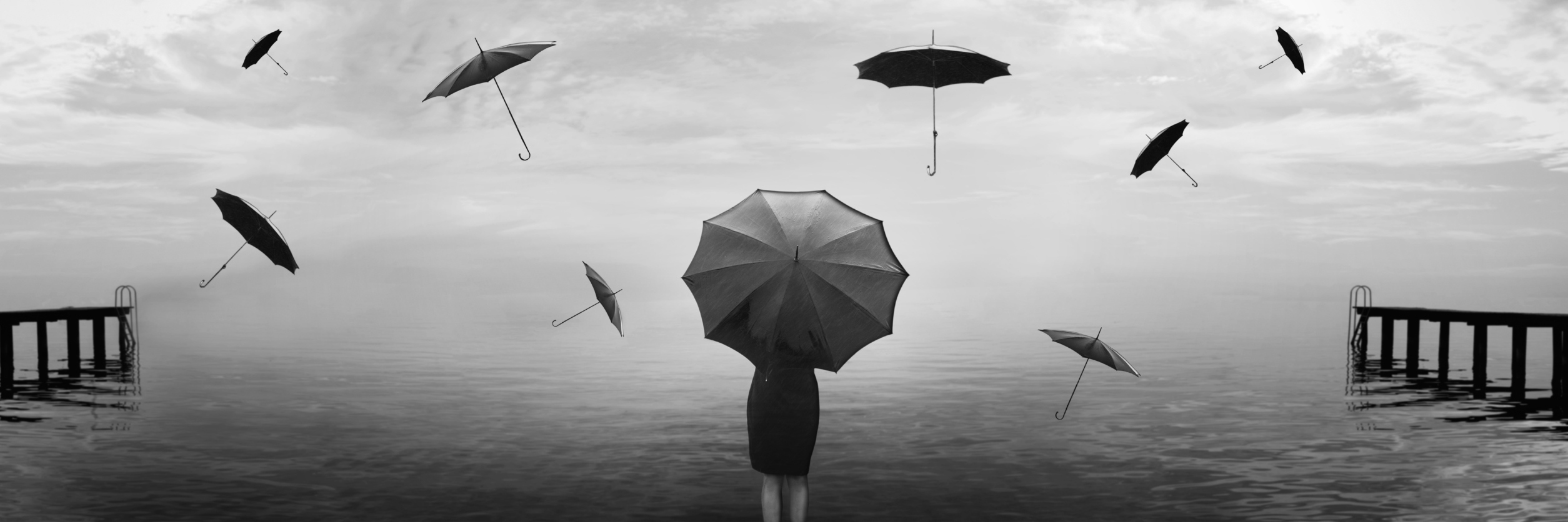 A woman stands at the end of the pier with umbrellas flying around her in the air.