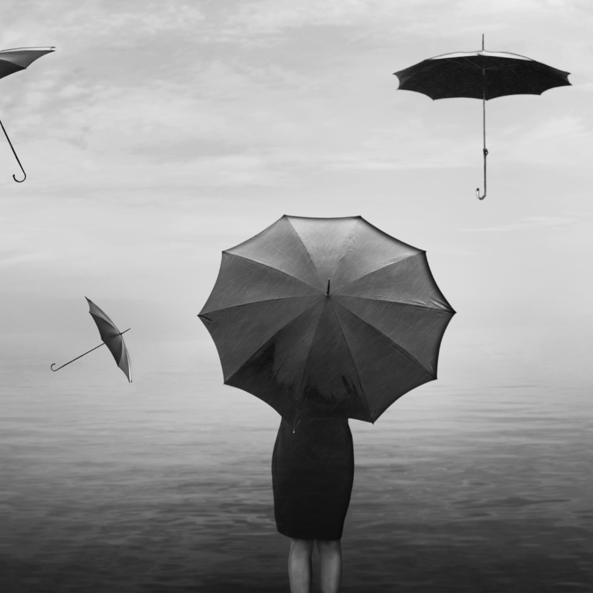 A woman stands at the end of the pier with umbrellas flying around her in the air.
