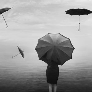 A woman stands at the end of the pier with umbrellas flying around her in the air.