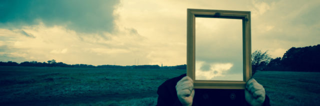 a man in a field holding a mirror over his face reflecting back clouds