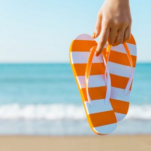 Woman's hand holding a pair of orange sandals against the coastline