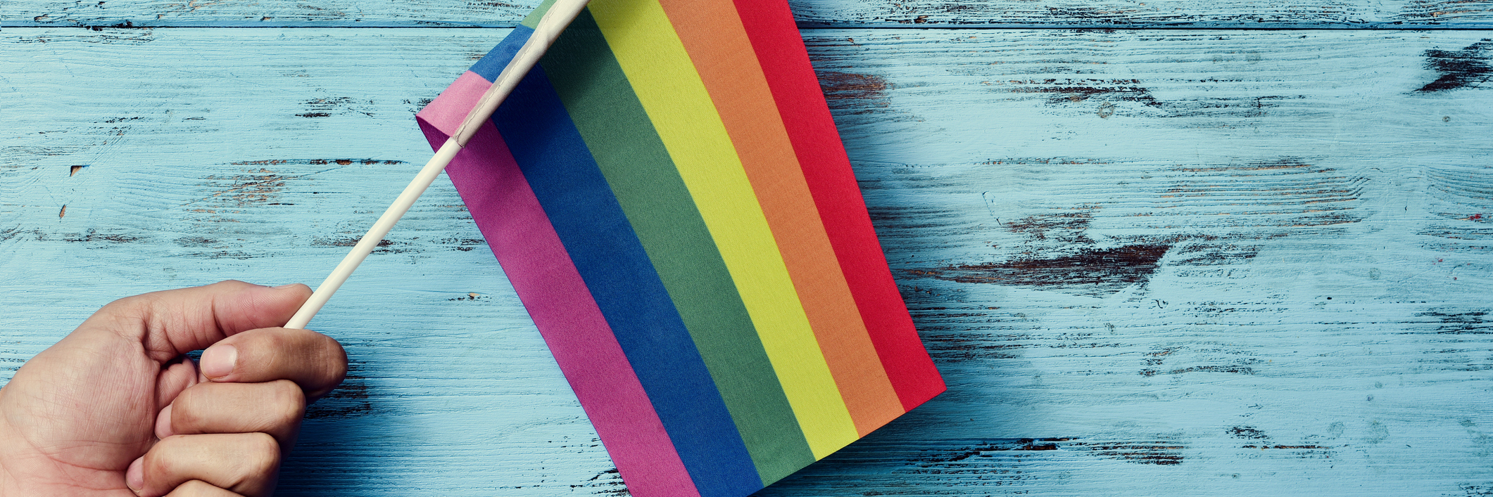 Why It's Difficult Attending a Pride Parade With a Chronic Illness Someone holding a rainbow flag against a blue background.