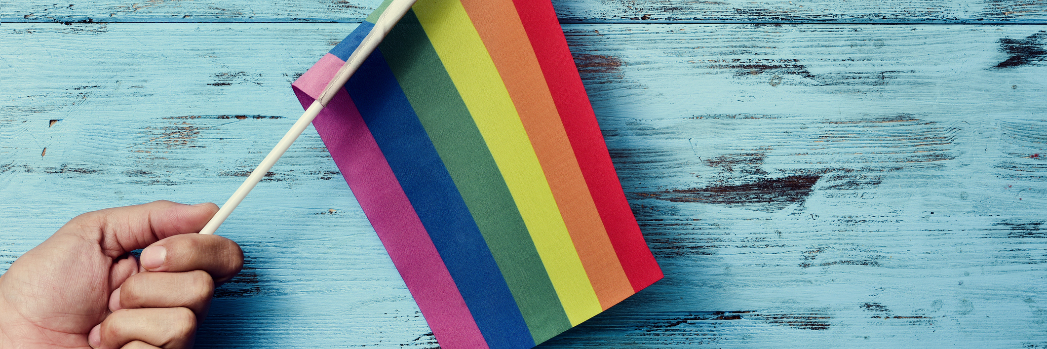 How Coming Out as Bisexual Helped Me Heal From My PTSD closeup of a young caucasian man waving a small rainbow flag against a rustic blue wooden background
