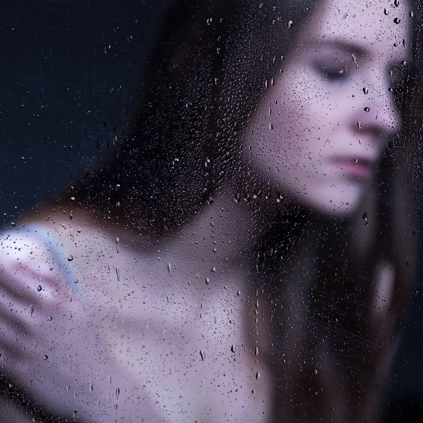 Woman stands behind glass with drops of water covering it.