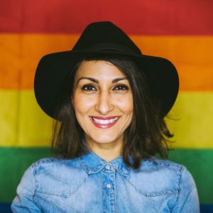A woman wearing a cowboy hat smiling in front of a rainbow gay pride flag