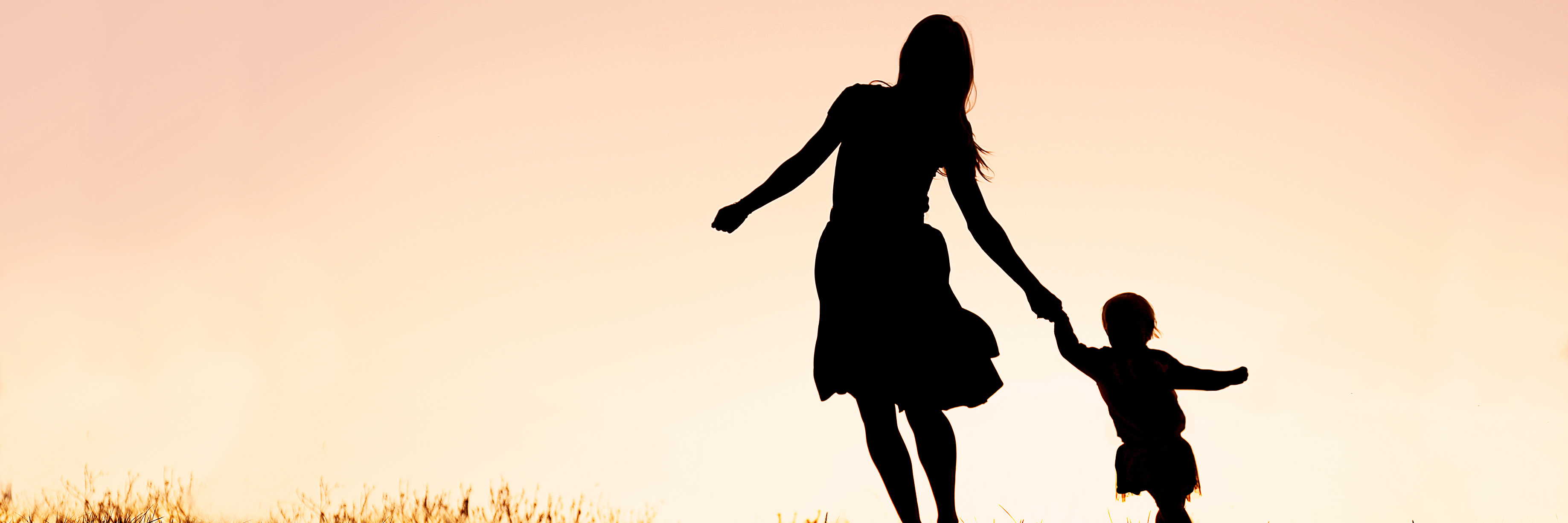 Silhouette of a happy mother and her baby daughter holding hands, and running and dancing outside at sunset.