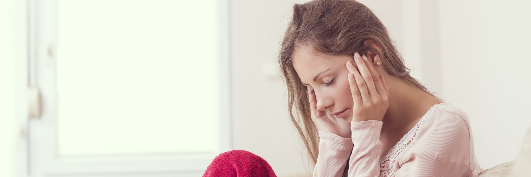 How You Know You Have a Chronic Illness Young woman sitting on a couch, holding her head, having a strong headache. Lens flare effect on the window