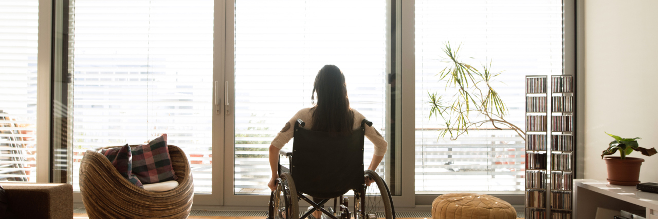 Young disabled woman in wheelchair at the window at home in a living room.
