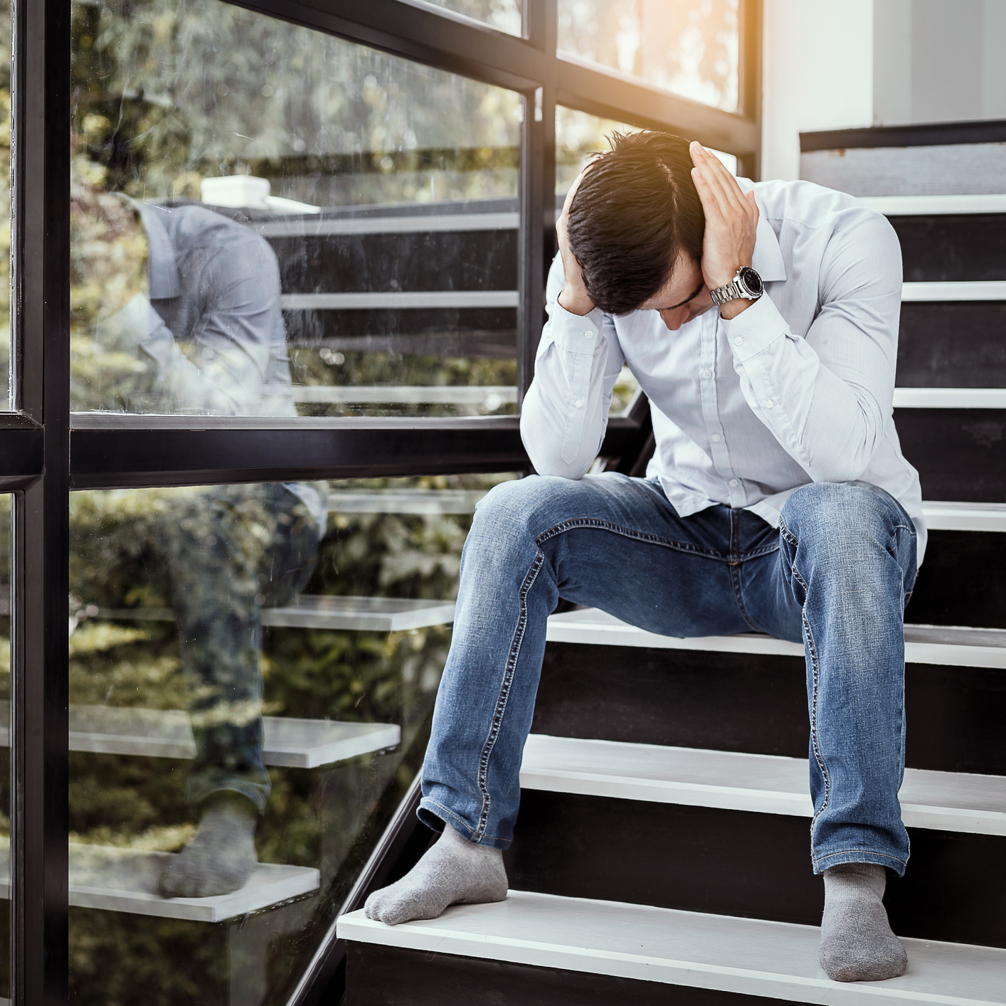 young man sitting on staircase near window holding head in despair or anxiety