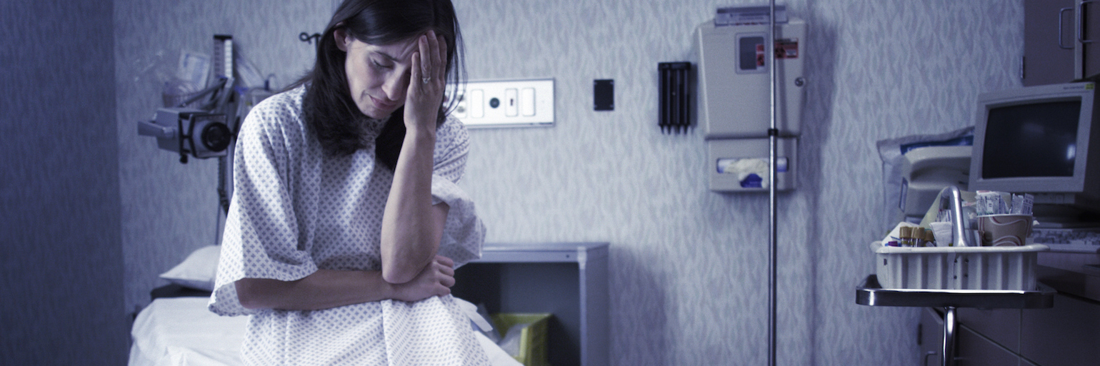 Female patient in hospital gown sitting in doctor's exam room, with hand over forehead, looking down