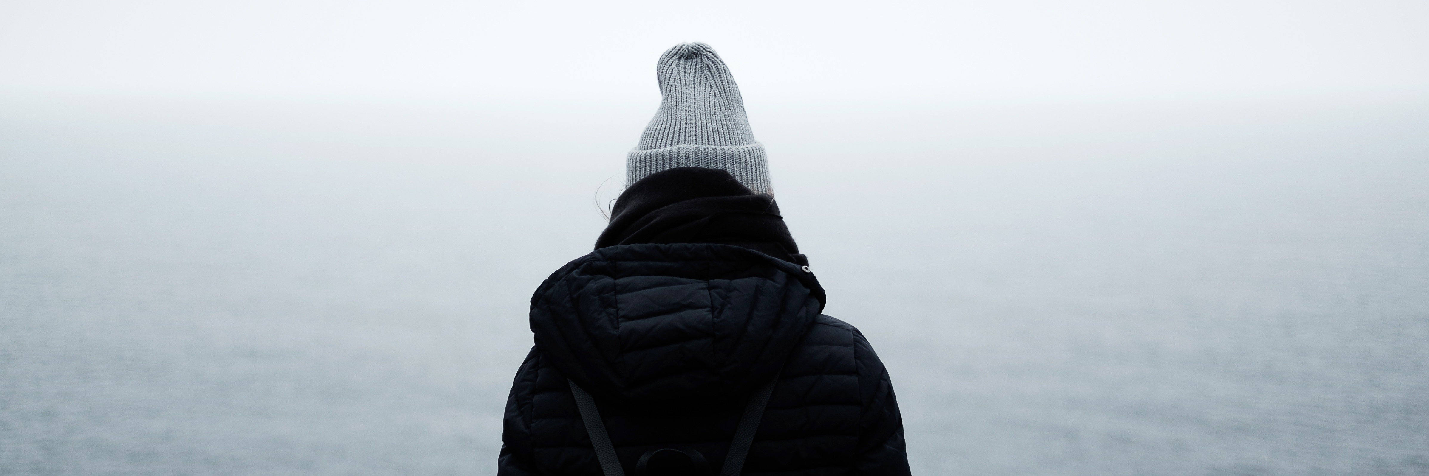 person looking out at misty ocean wearing hat and coat