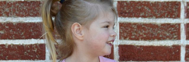 Picture of a little girl's profile, she has blond hair pulled in a ponytail, wearing a pink shirt and the background is a red brick wall.