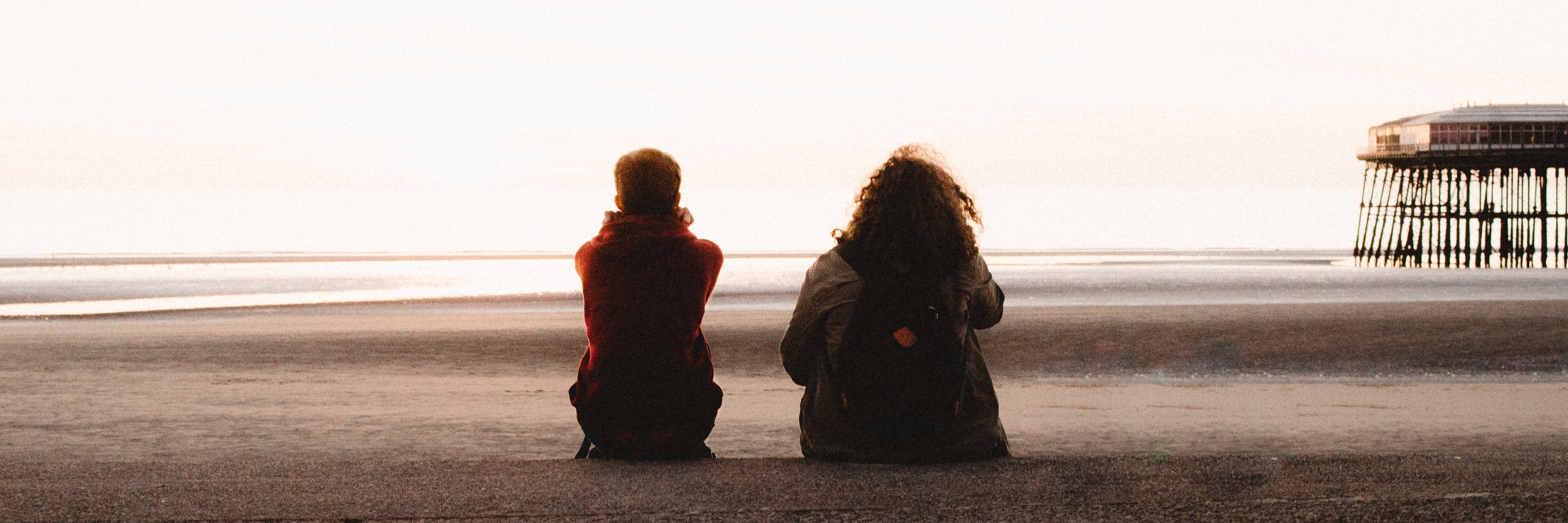 To My Friends Who Support Me Through My Mental Illness Two friends sitting on beach with backs to camera and pier in distance
