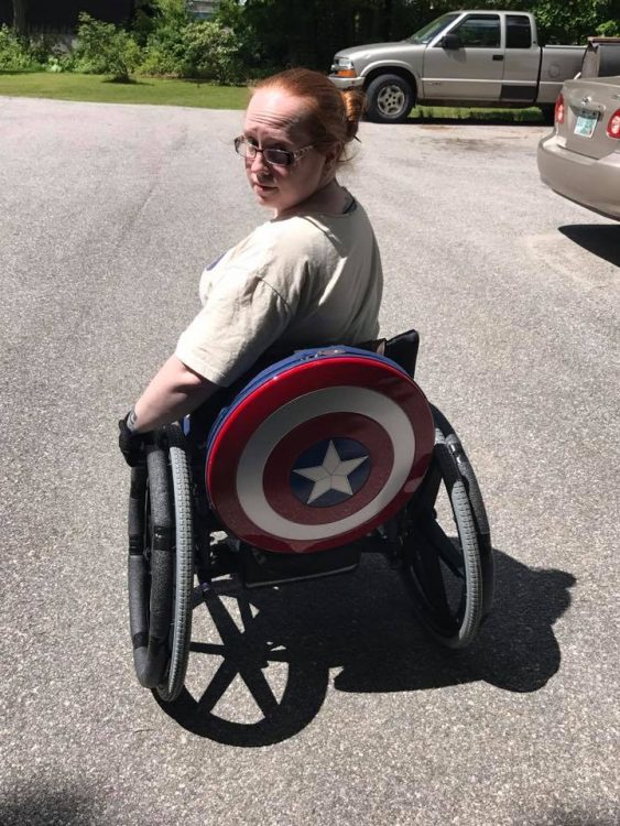 Woman sits in wheel chair, with a Captain America shield on back of her chair.