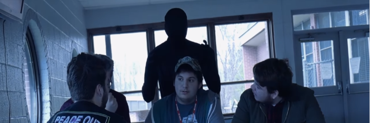 students at table with "anxiety"person in black morph suit