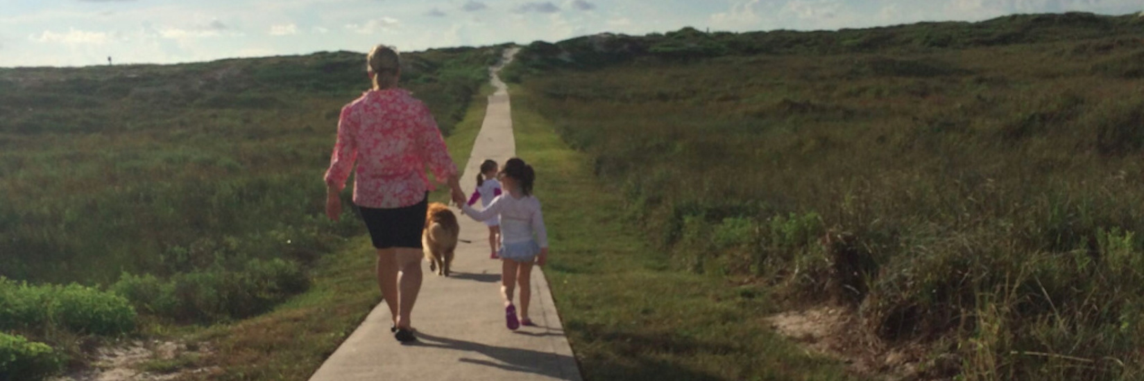 Mother holding daughter's or granddaughter's hand, walking with daughters or granddaughters down path surrounded by grass during the daytime