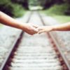 close up photo of two people's hands and arms standing either side of train tracks, reaching for each other