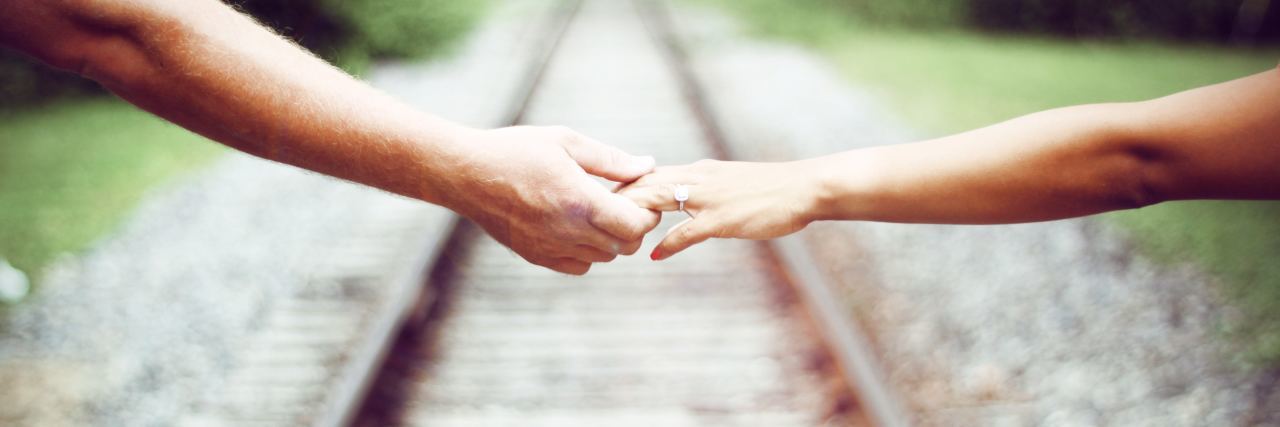 Wrong to Call Someone With Chronic Illness a 'Fighter' or 'Warrior' close up photo of two people's hands and arms standing either side of train tracks, reaching for each other