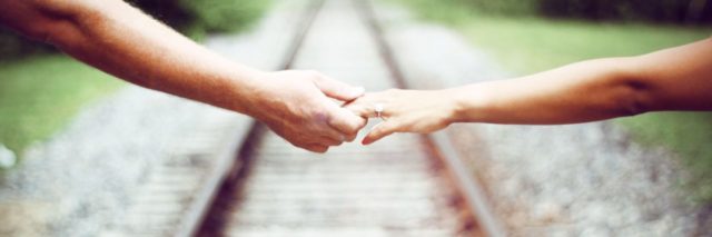 close up photo of two people's hands and arms standing either side of train tracks, reaching for each other