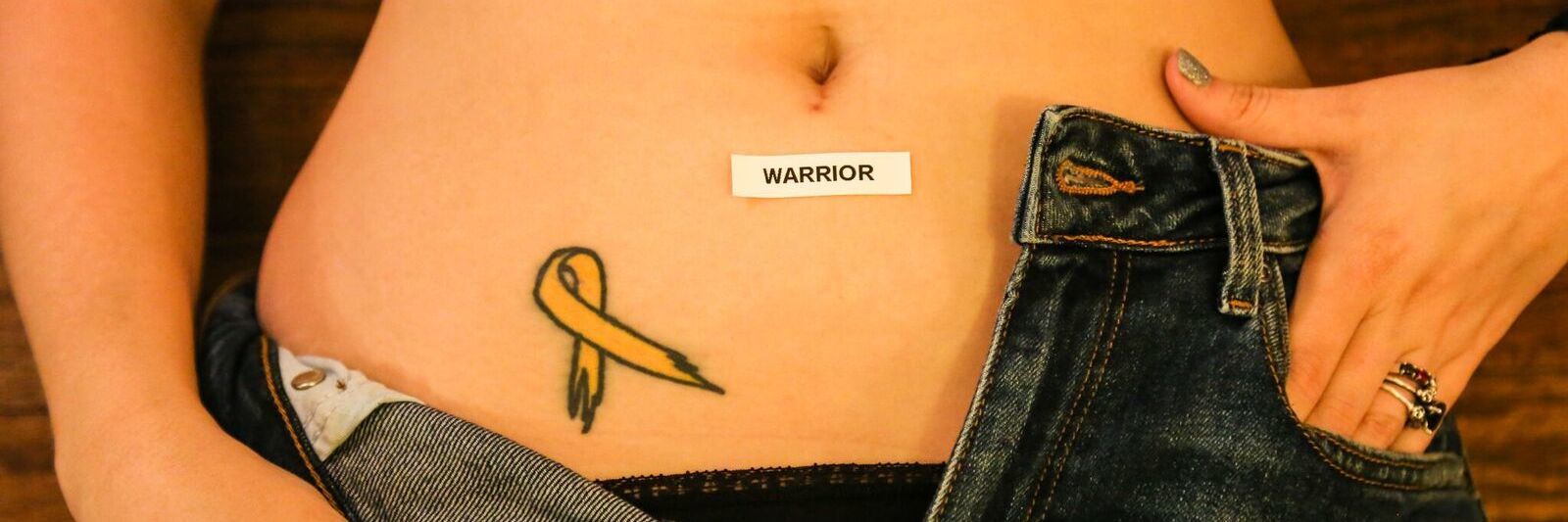 woman with her jeans unzipped to reveal her endometriosis awareness ribbon tattoo on her lower abdomen and a sticker on her stomach that says 'warrior'