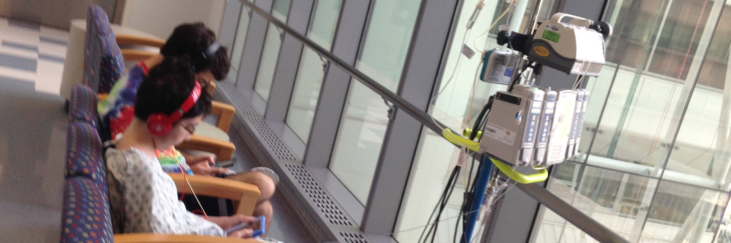 Coping and Self-Care When Your Child Is in the Hospital Two boys sitting in a hallway at a hospital, one boy has an IV and IV stand and hospital gown, they are playing video games