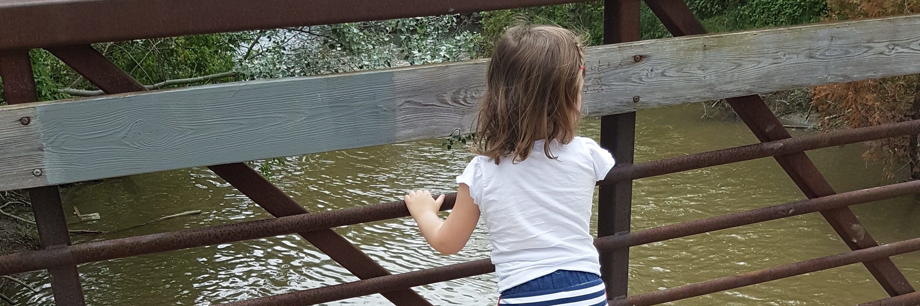 Little girl on a bridge looking at water. We see her back side and her leg braces.