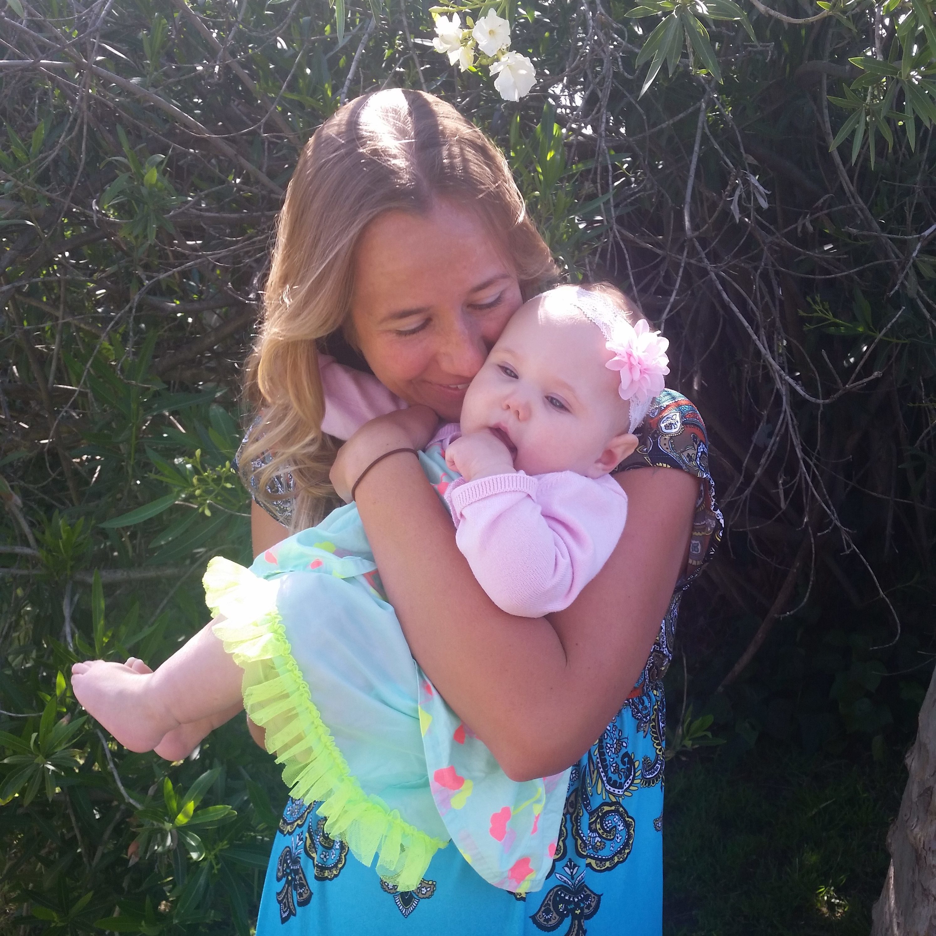 A mother wearing a blue dress holding her baby daughter who wears a pink sweater and teal dress.
