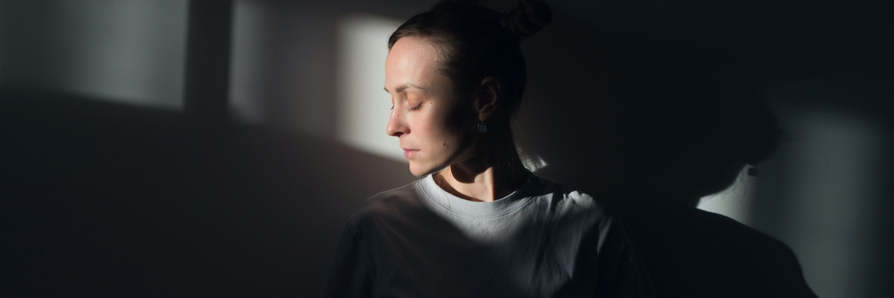 20 Surprising Physical Symptoms of Bipolar Disorder Woman standing in shadow with her face highlighted in rays of sunlight, with her eyes closed.
