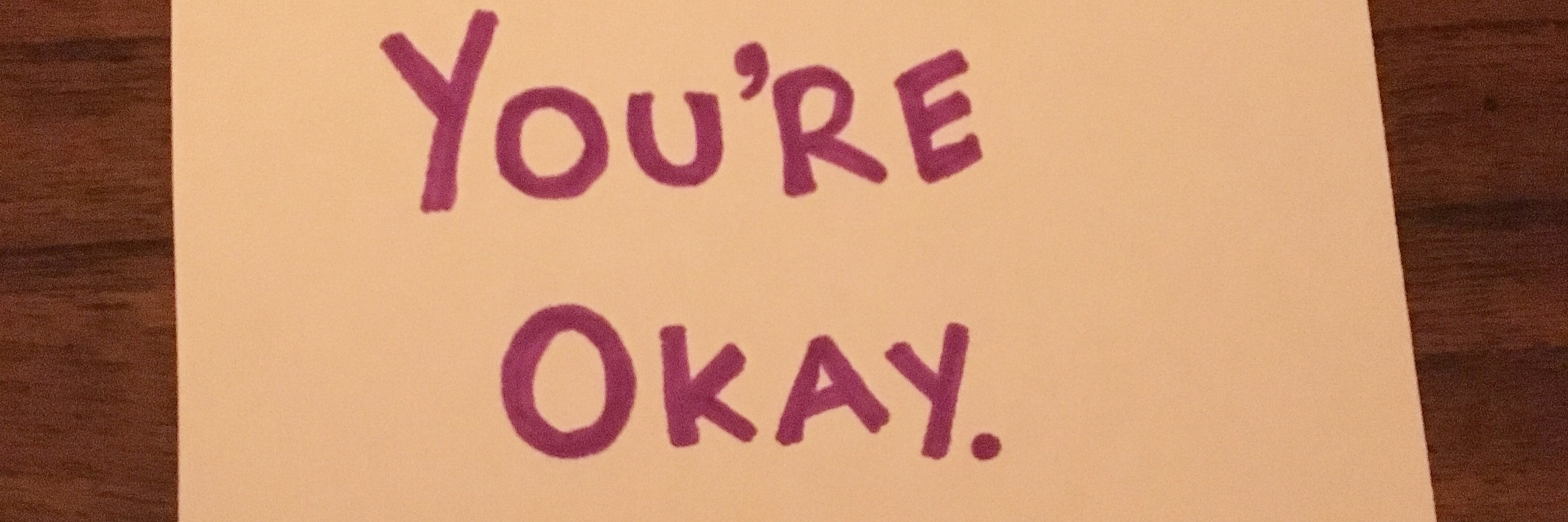 post it note on wooden table in pink text with words you're okay