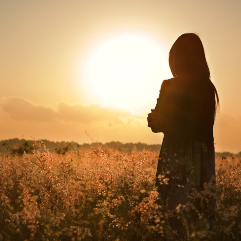 Woman standing in field, facing sun in sky