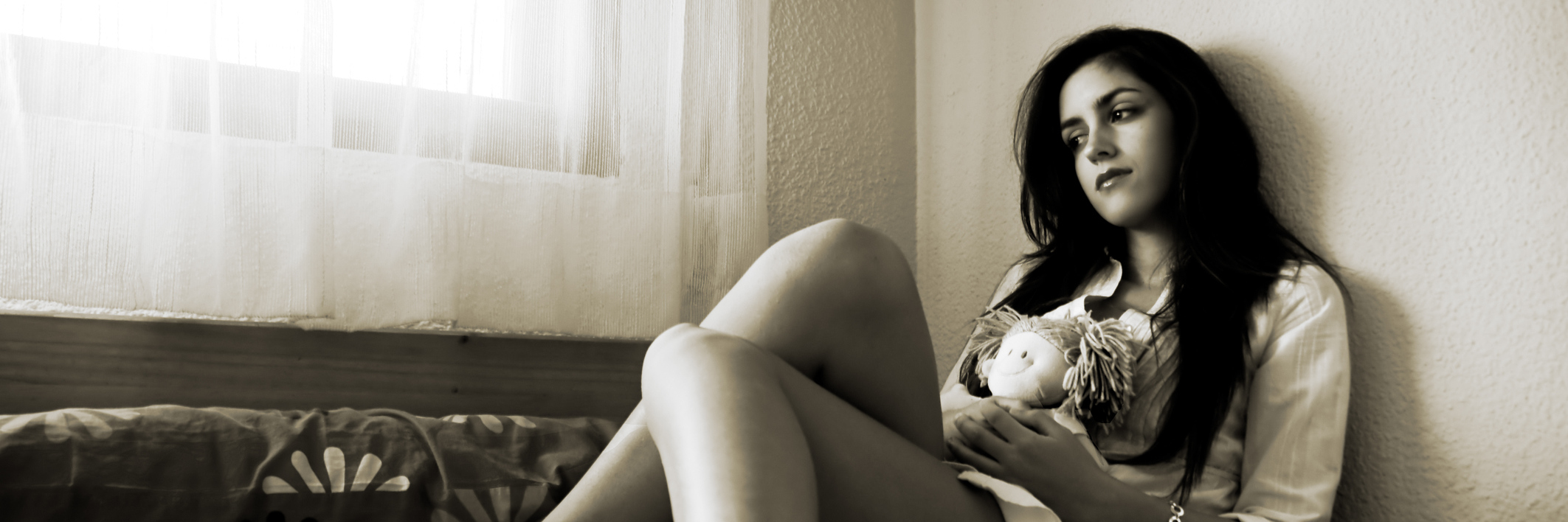 A teen girl sitting on her bed, looking out the window, with a solemn facial expression.