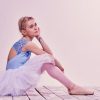 Tired ballet dancer sitting on the wooden floor, with a pink background behind her.