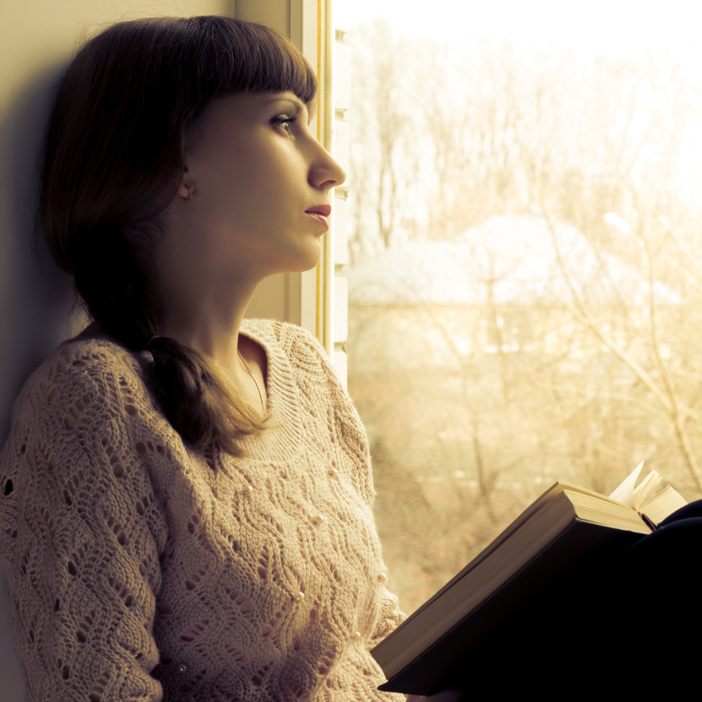 woman sitting next to a window reading a book and looking outside