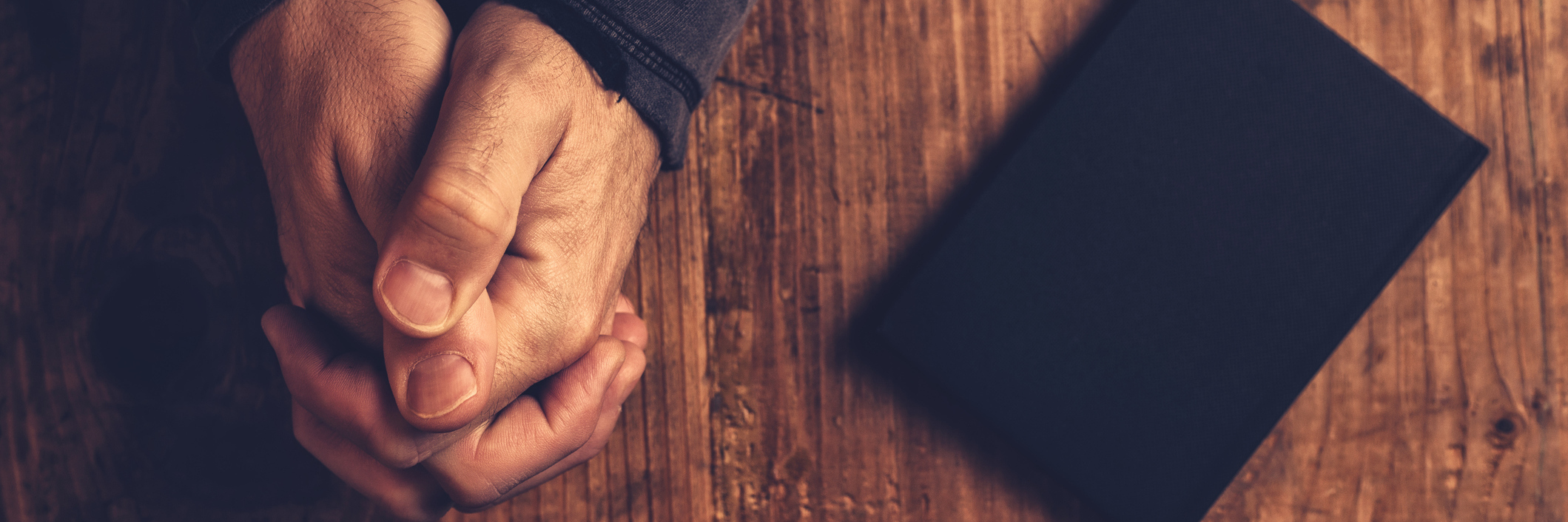 In Addition to Praying for Me While I'm Ill, Here's What You Can Do Hands of a Christian man on a wooden table, praying, with his Bible by his side.