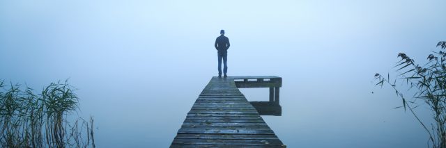 man standing alone on jetty over water on foggy day