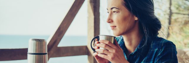 young woman sitting on wooden porch drinking coffee from thermos