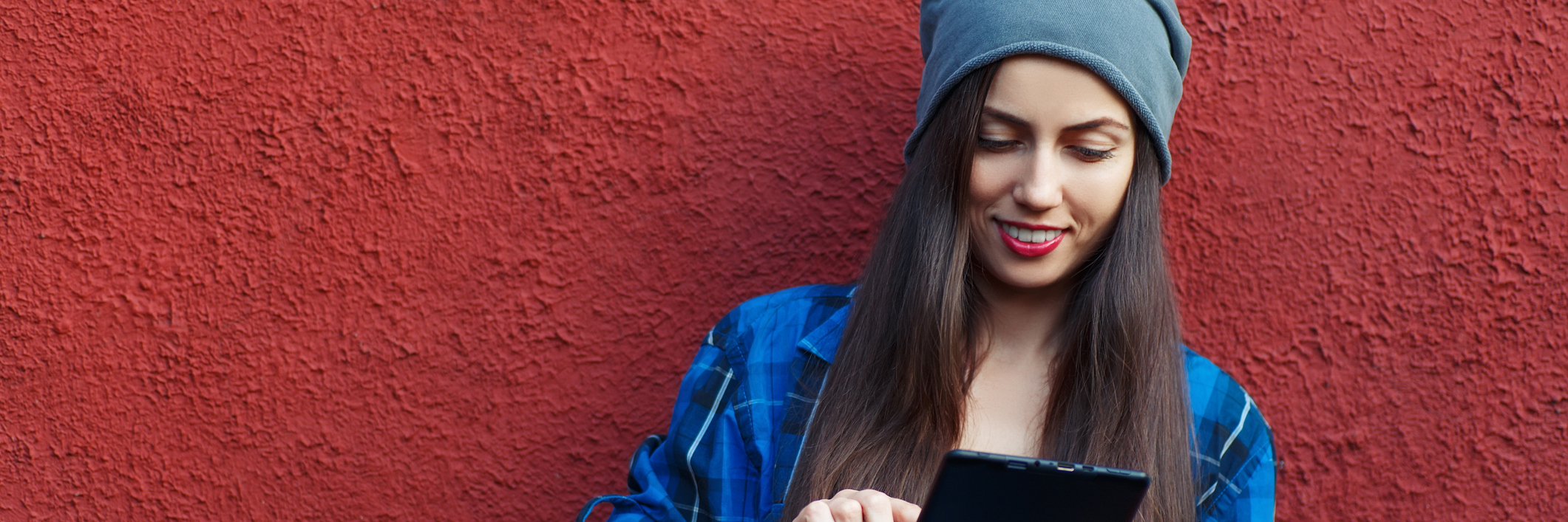 Portrait of a cheerful hipster woman holding tablet computer. Young hipster smiling girl using digital tablet outdoors, red wall in the background with copy space, social networking concept