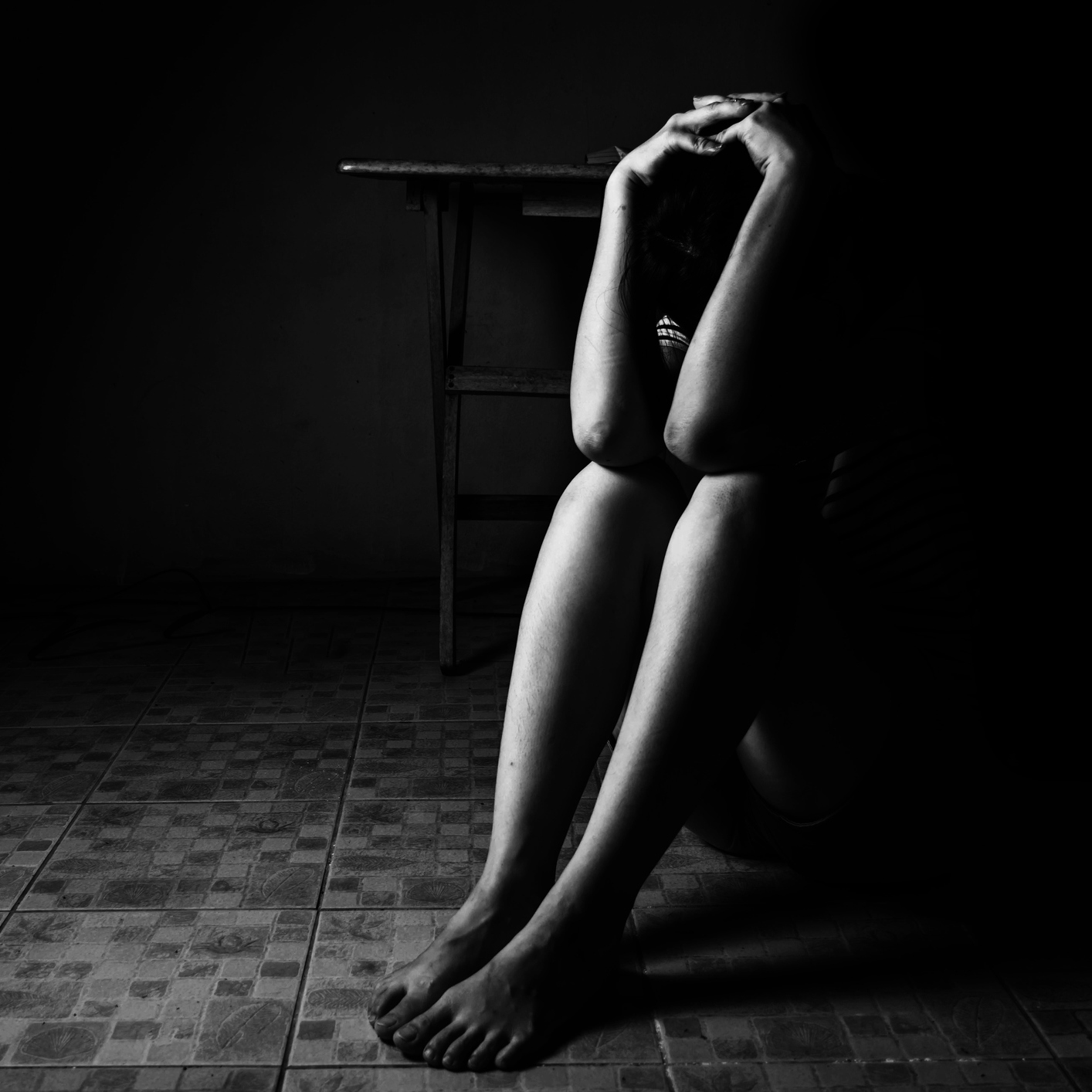 woman sitting on ground in darkness with arms raised to her head and elbows resting on knees