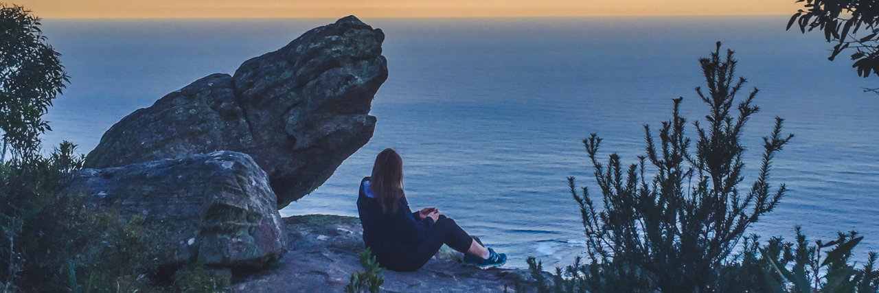 The writer sitting on some rocks, looking out towards a sunset and body of water.