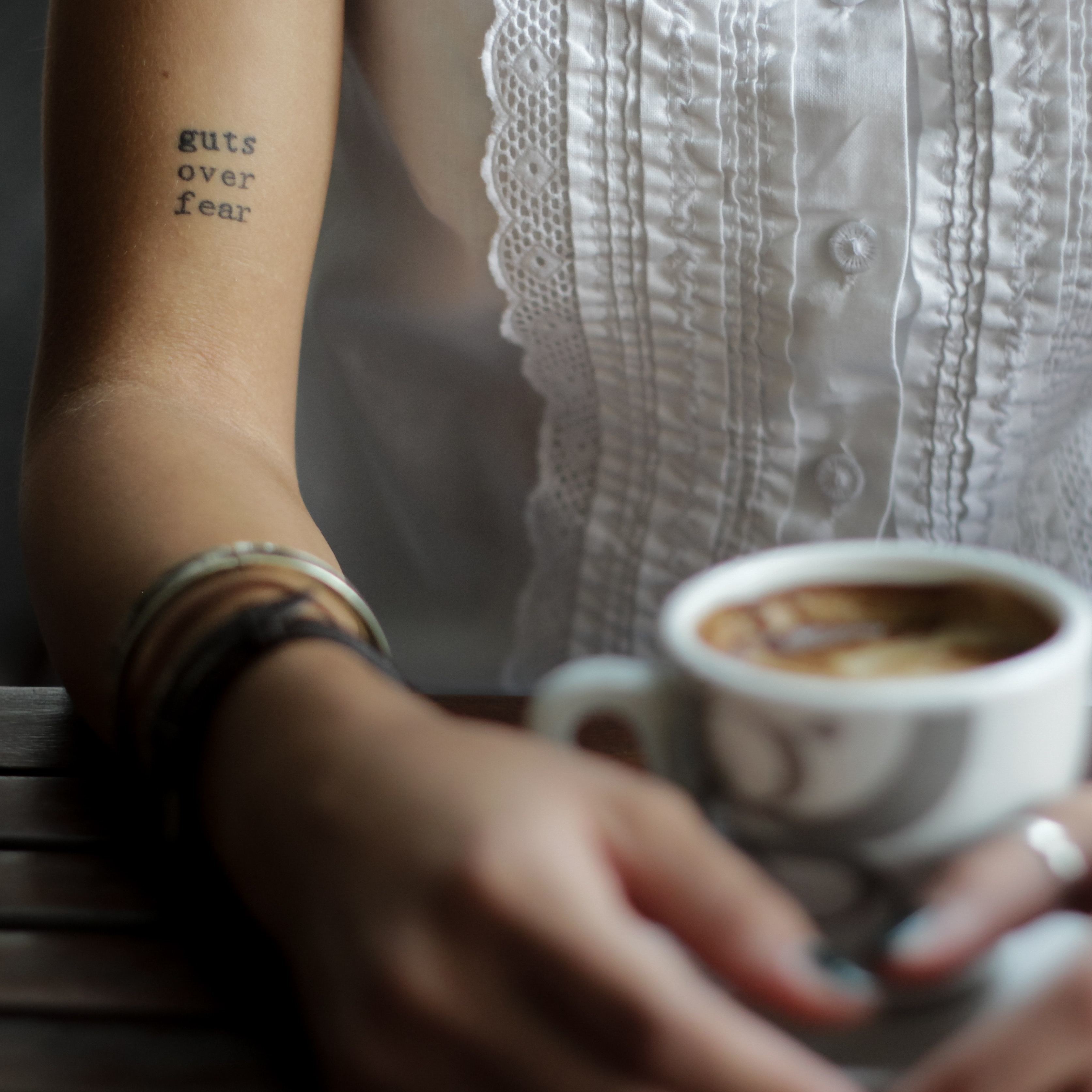 woman sitting at a table holding a coffee with a tattoo on her arm that says 'guts over fear'